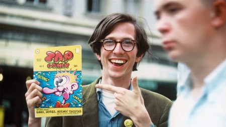 A smiling bespectacled man points to a comic book in his othe rhand. It shows a cartoon boy's face with crossed eyes, a giant nose, and a bow tie.