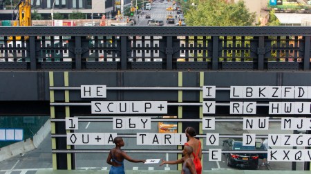 Three performers in jumpsuits stand on an overlook on the High Line arranging tiles of alphabetic characters in black text on white placards.