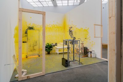 A room-within-a-room containing a bed, a peace lily, an exercise bike, a plexiglass shield, a vintage computer, and a paintball gun. It's all covered in yellow.