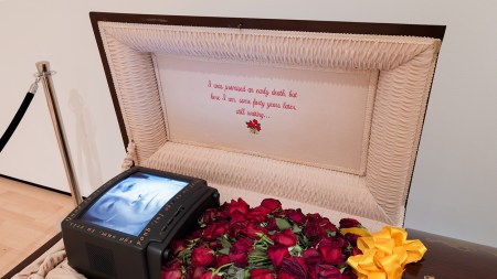A video monitor in a coffin shows a perseon's face with a breathing tube. The coffin is filled wit hwilting roses and a bottle of Jack Daniels is engraved on the outside. The inner lid is embroidered with the words: I was promised an early death, but here I am some forty years later, still waiting...