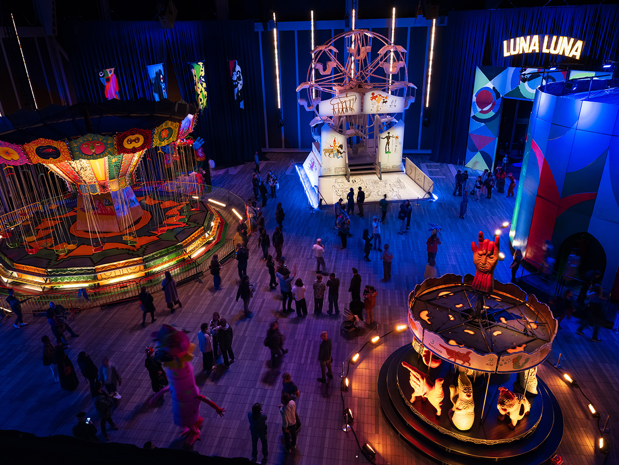 Aerial view of several amuseument park rides (carousels, swings, a merry-go-round) in a darkened interior space, lit with colorful lights.