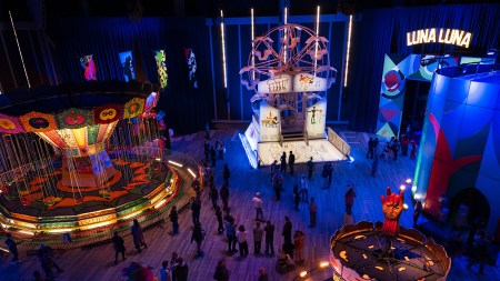 Aerial view of several amuseument park rides (carousels, swings, a merry-go-round) in a darkened interior space, lit with colorful lights.