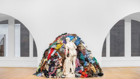Sculpture of a nude woman made with a white material. She is standing in contraposto and facing a pile of used clothes.