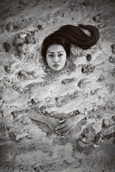 A black-and-white photograph of an Indigenous woman buried in cracked sand. Her face and hands are unburied. She stares at the viewer. 