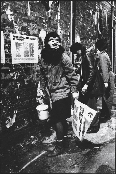 A black and white photograph of a person wearing a gorilla mask holding a bucket of glue in one hand and posters in the other hand. They are standing in front of a wall where a poster is on display. 