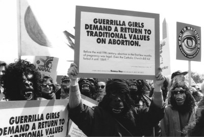 A black and white photograph of a group of people wearing gorilla masks and holding up signs. The person in the center holds a sign that reads “Guerrilla Girls demand a return to traditional values on abortion.”   