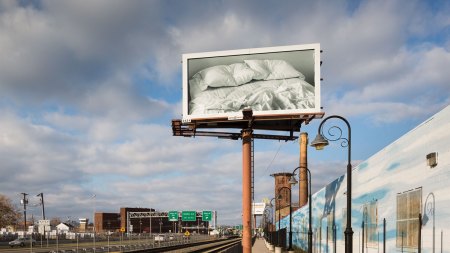 A photo a billboard of an empty bed with two pillows having an indention. It is installed by railroad tracks.