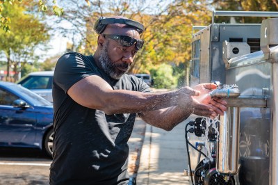 A Black mean is wearing Okaley's and headphoens on top of his head, cupping his hands to catch water spraying from a machine.