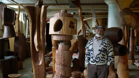 A Black man standing beside wood sculptures.
