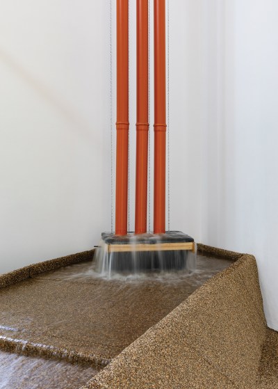 Three vertical red pipes funnel water down into a basin made of a brown, gritty material.