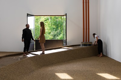 Inside a gallery, water flows through a basin made of a brown, gritty material in front of a window open to a lush verdant scene.