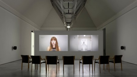 5 chairs face a wall in a gallery with a vaulted roof. Two video projections are shown side-by-side. One shows a white woman with long brown hair and bangs facing the camera in front of a gray wall. The other shows a long white institutional hallway.