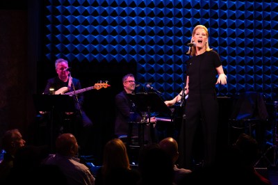 A blond woman singing at a microphone in front of a bassist and piano player.