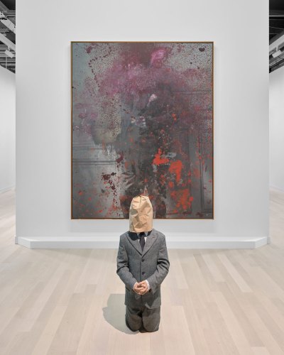 A sculpture of a kneeling man with clasped hands in a tweed suit with a bag over his face stands in front of an abstract painting with a specterly figure. 