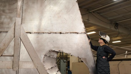 A white woman in a black padded jacket and dark pants stands with hands on hips in front of a large, metallic, geometric-shaped sculpture in an industrial workshop setting. The sculpture has rough surfaces and an open circular area. The environment includes visible machinery and the ceiling is supported by beams.