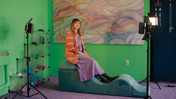 A white woman in an orange tunic and purple dress sits on an undulating hunk of foam in a green room.