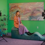 A white woman in an orange tunic and purple dress sits on an undulating hunk of foam in a green room.