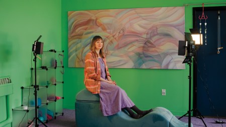A white woman in an orange tunic and purple dress sits on an undulating hunk of foam in a green room.