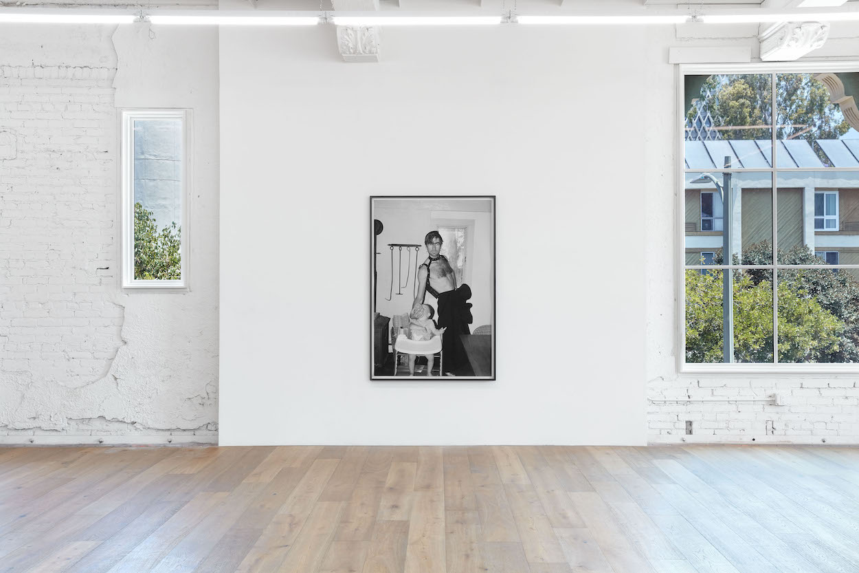 Within a large windowed gallery space hangs a framed photograph depicting a man standing topless in a skirt and feeding a baby in a highchair.