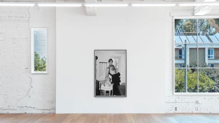 Within a large windowed gallery space hangs a framed photograph depicting a man standing topless in a skirt and feeding a baby in a highchair.