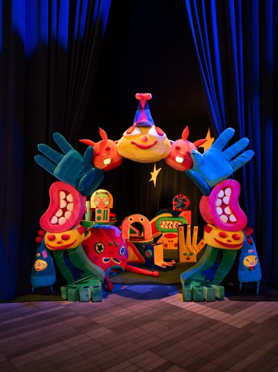 A round archway comprises a smiley face, hands, two mouths, and legs. It is colorful and soft, and gives way to a portal brimming with plushies.
