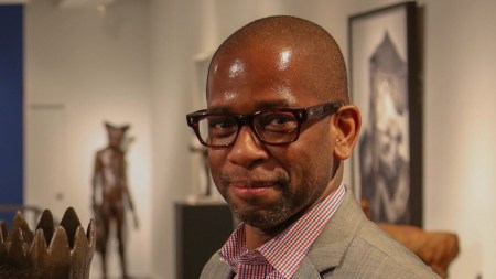 A black man with black rectangular glasses smiles at the camera in a gray suit and checkered shirt.