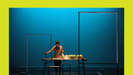 In a black box space, a black person slices watermelons on a table.