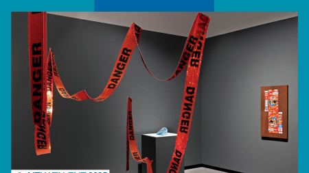 A grey-walled gallery with orange beaded orange "Danger" tape hanging from teh ceiling.