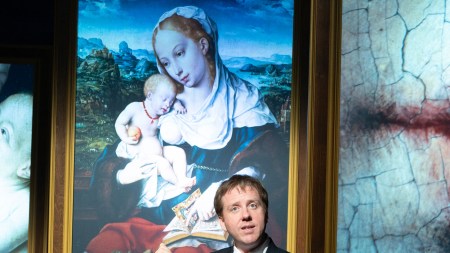A white man in a suit and tie points to a madonna and child painting projected behind him.