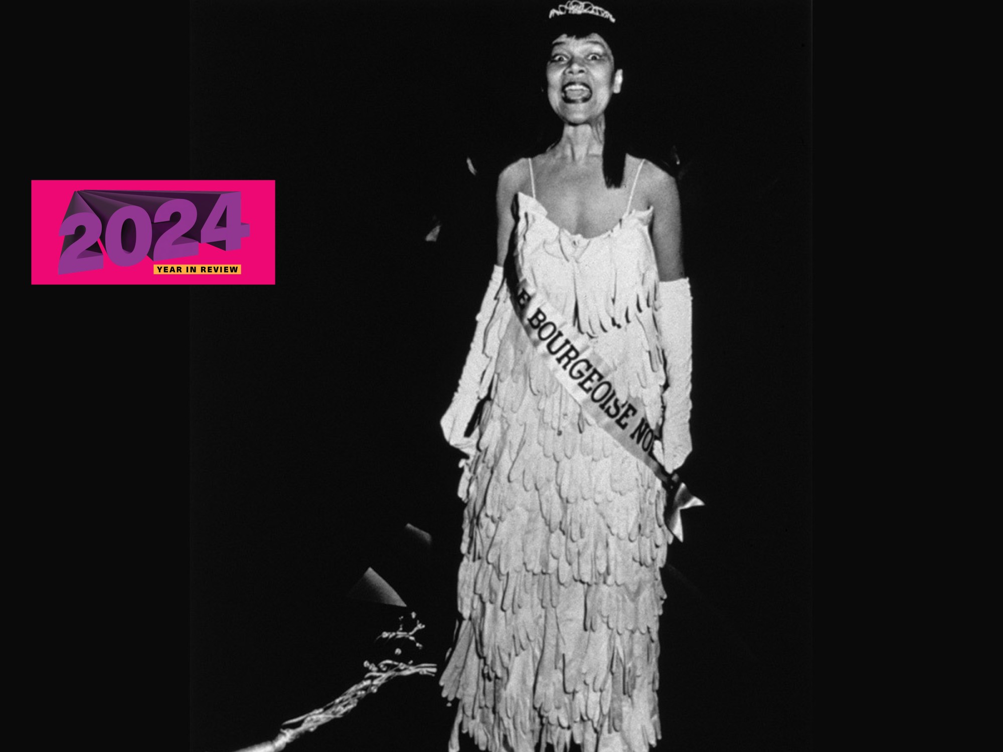 A Black woman wearing a dress made out of gloves with a sash reading 'BOURGEOISE' running across it. She shouts before a black background.