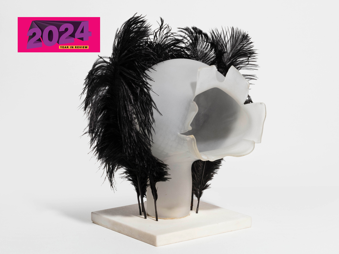A glass sculpture of an abstracted head with a face blown open to reveal a hollow inside. Black feathers surround the head.