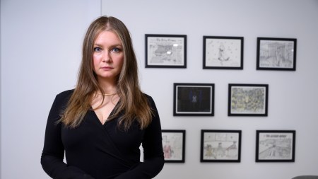 A woman wearing a black dress looks at the camera in front of eight framed drawings.