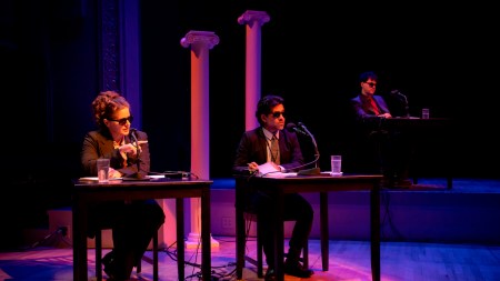 Three people dressed as interrogators sit at tables on a stage lit with orange, blue, and purple hues.