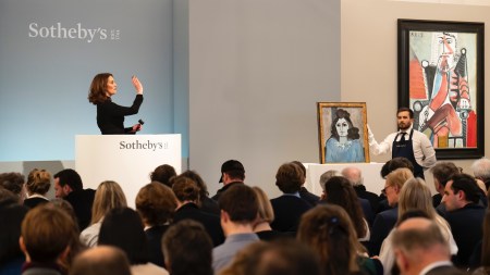 A woman standing behind a dais gestures at a crowd beside two paintings.