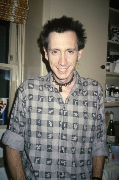 A somewhat femme white man with a spiked collar, floofed hair, and single dangling earring. He's wearing a gray gingham shirt with animal sillhouettes in the darkest squares.