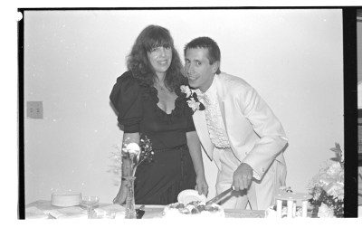 A man and a woman cutting a cake, possibly at their wedding. She is wearing a black dess with poofy sleeves, and he is wearing a white suit with a frilly shirt, bending over.