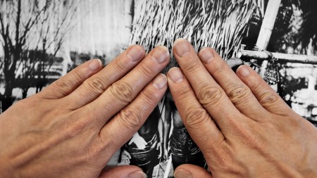 Two olive skinned hands cover figures in a black-and-white photograph. The figures are barefoot in the sand; a bamboo structure is visible in the background.