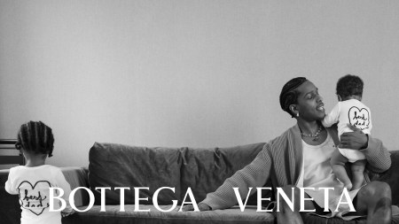 A$AP Rocky, a Black man, sits on the couch holding a baby. A toddler is standing facing the other end of the couch. This is a square black-and-white photo and Bottega Veneta is printed over it.