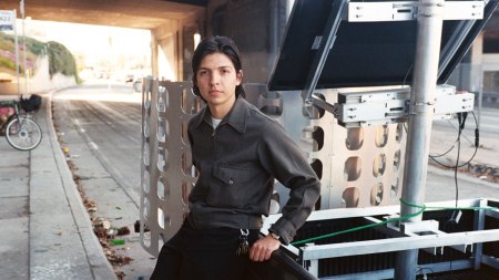 Portrait of Carlos Agredano leaning against a pickup truck near a freeway overpass.