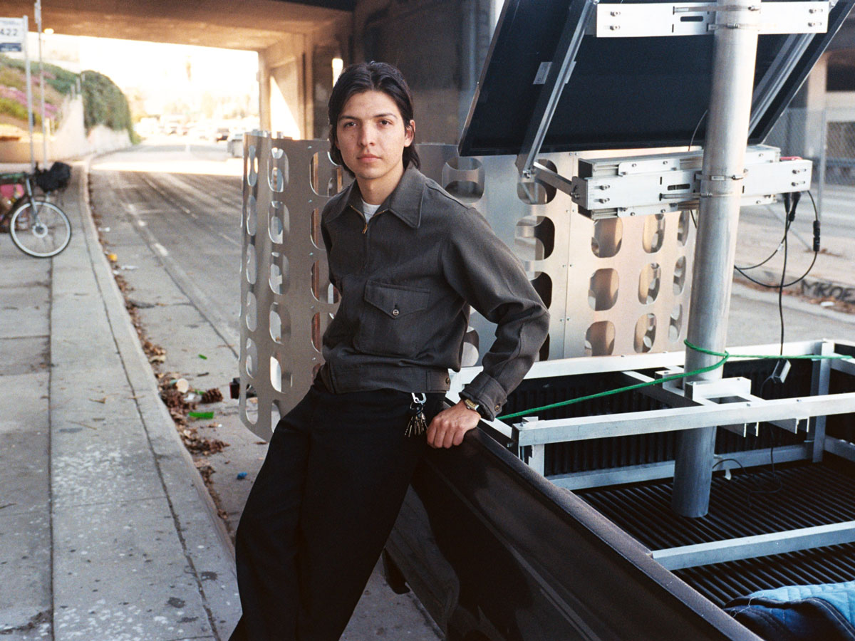 Portrait of Carlos Agredano leaning against a pickup truck near a freeway overpass.