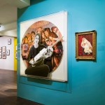 An installation shot of a gallery with teal and yellow walls, and a large circular installation with a woman cradling an object in front of a group of other figures.