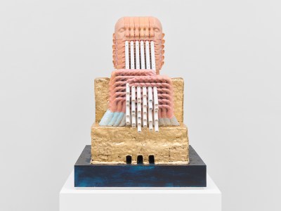 A bust of a bald person playing a flute is repeated 36 times and amalgamted into a kind of grid, as if forming one, and not 36 humans. The person's arms and head emerge from a golden stepped form rather than a body.