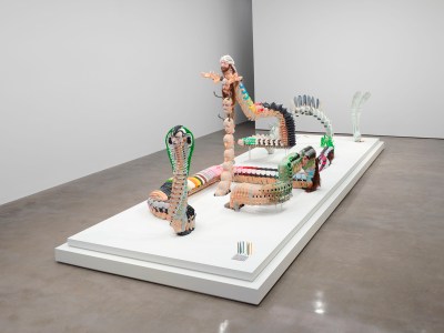 On a low plinth, an undulating serpent made of many colorful heads, with a bunny head peeking out in the back.