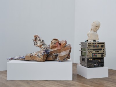 A sculpture made of two elements. One is a clay form covered in newspaper clippings and exhibited on a plinth. The other is a clay bust on a stack of video recorders.