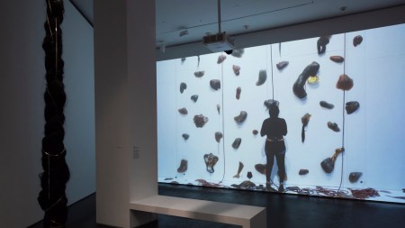 Within a darkened gallery, a projection shows a person standing beneath what appears to be a rock climbing wall. From the ceiling nearby hangs a thick, long braid resembling a rope.