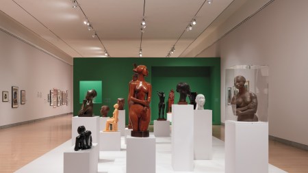 A plinth is littered with a dozen or so figurative sculptures, busts and full size. Most appear to be of women, and are of blocky figures carved in wood and stone. A green wall is behind them.
