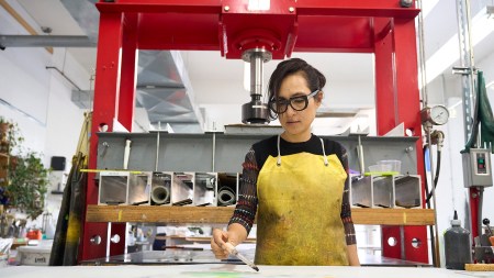 A woman with dark hair and dark glasses applies colored liquid to a work on paper.