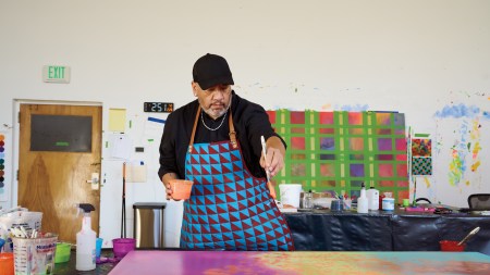 A man in a patterned painter's smock dripping paint onto a canvas from a brush held aloft.