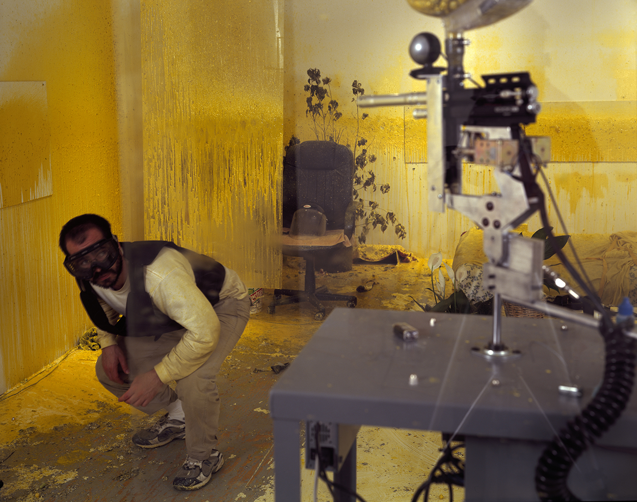 A brown man with ski goggles on dodges a paintball gun in a room covered in yellow paint.