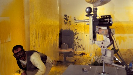 A brown man with ski goggles on dodges a paintball gun in a room covered in yellow paint.